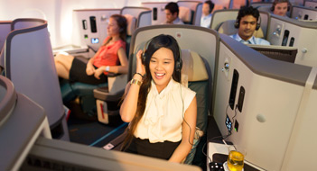 Female in business class wearing headphones looking at the screen in front of her with an open-mouthed smile