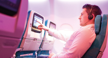 A male passenger with headphones on reclines in seat pressing a button on the entertainment system in front of him