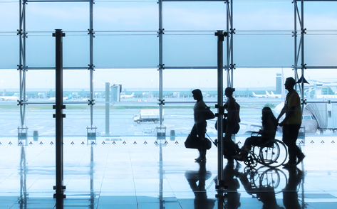 Silhouette of four individuals in an airport terminal overlooking an Airport parking bay. One individual is pushing a wheelchair